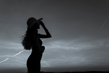A woman wearing a hat and standing in the sunset. She is reaching out with her arms open, as if she...