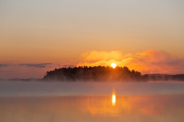 Sun rising over an island on a calm lake on a foggy morning