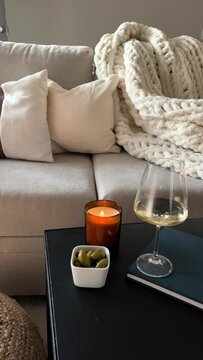 Female hand putting burning candle near the glass of white wine. Cozy atmosphere at home, beige tones living room