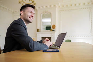 Smiling businessman at an elegant wooden table with a laptop and smartphone in a luxurious classical interior.