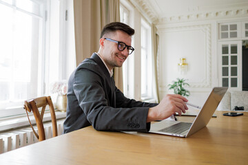 Fototapeta premium Focused executive in a suit works on a laptop in a bright, classic office, embodying professionalism and dedication.