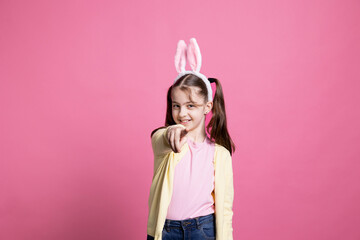 Obraz na płótnie Canvas Young schoolgirl with pigtails dancing around in pink studio, pointing at something in front of the camera and having fun. Joyful small child feeling confident with bunny ears for easter.