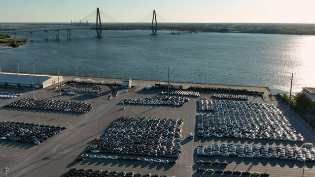 New cars parked in vast lots with Arthur Ravenel Jr. Bridge in the background at sunrise.