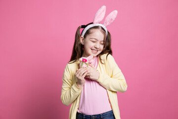 Obraz na płótnie Canvas Happy excited young girl posing with a stuffed rabbit in studio, wearing bunny ears and holding easter decoration in front of the camera. Young child with pigtails being proud of fluffy toy.