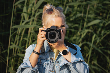 A blonde girl photographer looks through the viewfinder of a photo camera in nature.