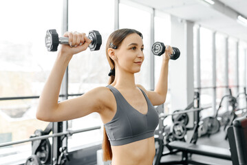 young athletic caucasian woman trains in fitness gym, standing and doing dumbbell press up,...