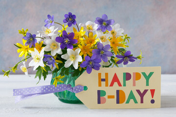Bouquet of spring blue, purple, white and yellow flowers in a vase on the table and a card with the text happy birthday. - 782509905