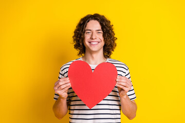 Photo of nice young man hold heart shape card wear striped t-shirt isolated on yellow color...