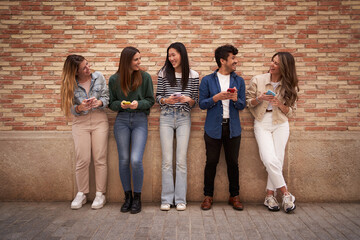 Group diverse happy young friends leaning on brick wall looking at each others holding mobiles...