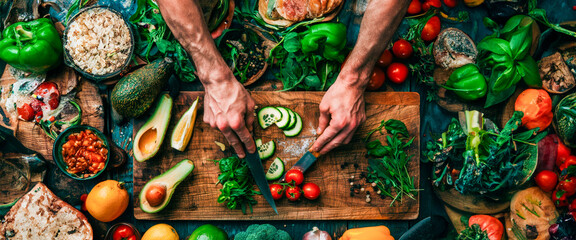Overhead view of hands chopping fresh vegetables on a wooden board surrounded by a rich assortment of colorful, healthy ingredients and homemade dishes. Banner. Copy space