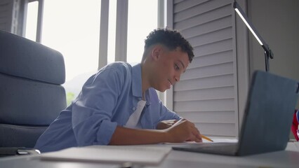 Teenage boy sitting at desk in bedroom at home doing homework on laptop - shot in real time - Powered by Adobe