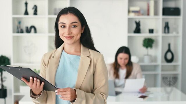 Positive friendly successful beautiful indian or arabian business woman, secretary, company executive, in a stylish suit stand in a modern office, holding documents, looking and smiles at camera