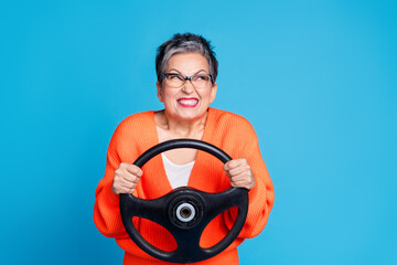 Photo portrait of senior lady hold steering wheel look tricky empty space dressed stylish orange garment isolated on blue color background