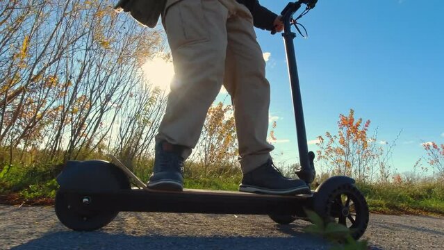 Riding electric scooter through the green area of the city park. Asphalt road and sun set. Evening recreational leisure. Driving ecological environmentally conscious power transport. Active lifestyle.