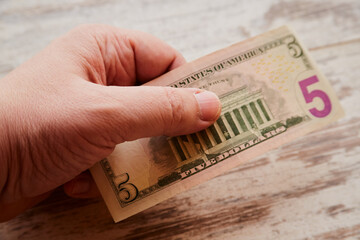 A hand holding five dollars against a worn wooden background, poverty and low wage