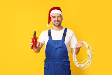 Portrait of male electrician in Santa hat with pliers and wires on yellow background