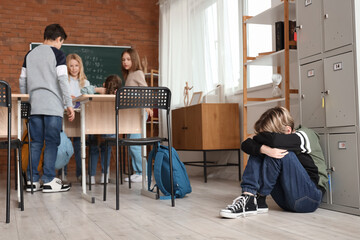 Bullied little boy sitting near locker in classroom - obrazy, fototapety, plakaty