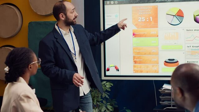 Management consultant gathering his employees for a status update meeting in boardroom, interactive presentation with the latest data. Success metrics and key performance indicators. Camera A.