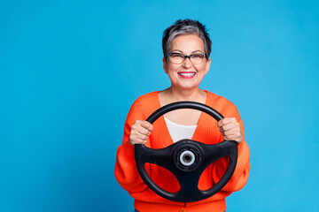 Photo portrait of lovely senior lady hold steering wheel dressed stylish orange garment isolated on blue color background - obrazy, fototapety, plakaty