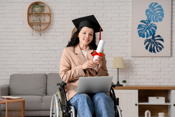 Beautiful female graduate student in wheelchair with graduation cap, diploma and modern laptop at...