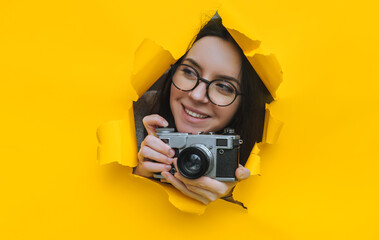 A paparazzi girl in glasses with a rare SLR camera looks out from her hiding place and smiles while watching what is happening.Yellow paper, torn hole.Tabloid press.Looking for a subject, stock photo. - obrazy, fototapety, plakaty