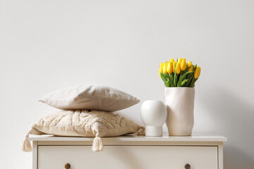 Vases with tulips and pillows on commode near light wall