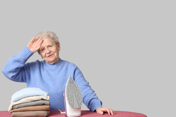 Tired senior woman with stack of laundry and iron on grey background - Powered by Adobe