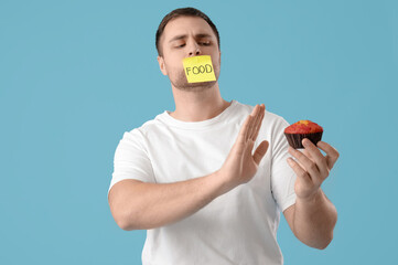 Young man with taped mouth rejecting muffin on blue background. Weight loss concept
