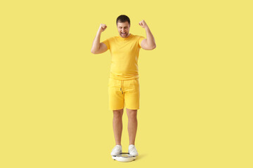 Happy young man measuring his weight on scales against yellow background