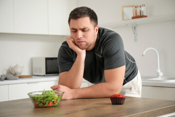 Fototapeta na wymiar Sporty young man with salad and muffin on table in kitchen. Weight loss concept
