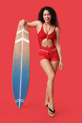 Beautiful young happy African-American female lifeguard with surfboard on red background