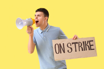 Protesting young man holding placard with text ON STRIKE and shouting into megaphone against yellow...