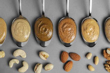 Tasty nut butters in spoons and raw nuts on gray table, flat lay