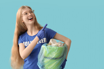 Female volunteer holding recycle bin with paper pointing at something on blue background - obrazy, fototapety, plakaty