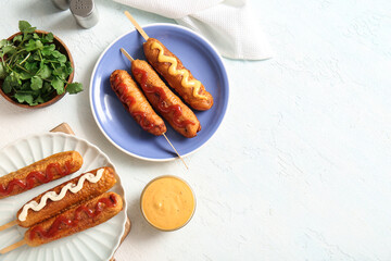 Plates of tasty corn dogs with different sauces on white background