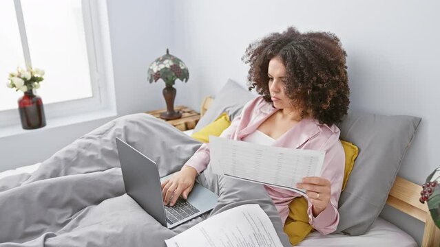 A young woman with curly hair works on her laptop in bed, surrounded by papers in a cozy bedroom setting.