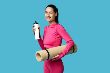 Young sporty woman with water bottle and yoga mat on blue background