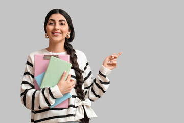 Beautiful young happy Indian woman with notebooks pointing at something on grey background