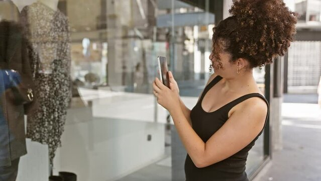 Young woman with curly hair using smartphone on a city street, reflecting urban shopping scene.
