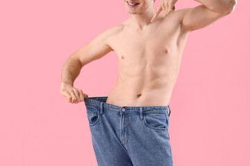 Handsome happy young sporty man pointing at loose jeans on pink background, closeup. Weight loss concept