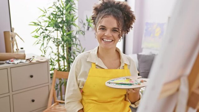 Cheerful hispanic artist woman with curly hair flaunts dazzling dental health, pointing at her teeth, happily sitting in art studio