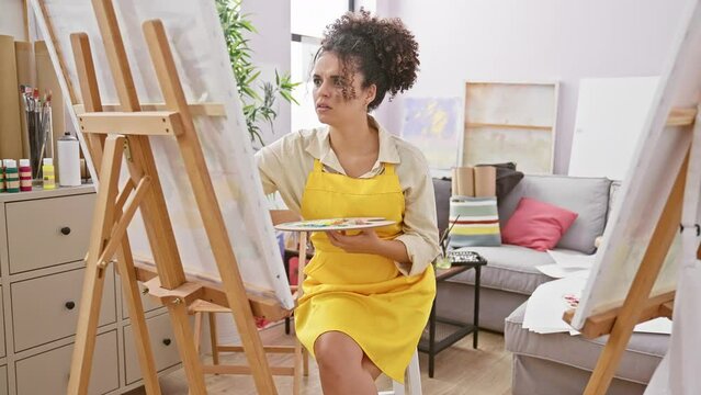 Eccentric hispanic woman artist with curly hair, sitting in her studio, hands on head, sporting a crazy surprised look, scared out of her wits, mouth agape in a state of shock!