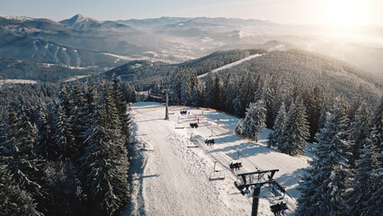 Sun ski resort at snow mountain forest aerial. Escalator chair at pine trees. Tourist attraction. Active sport. Winter vacation at Carpathians mounts, Bukovel, Ukraine, Europe. Travel, tourism scenery - obrazy, fototapety, plakaty
