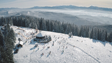 Winter active sport resort at snow mountain top aerial. People at nature landscape. Ski slope at fir forest. Mount ranges with pine trees. Tourist attraction at Carpathians, Bukovel, Ukraine, Europe