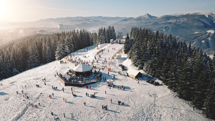 Sun ski resort aerial. Snow mountain top. Nature landscape. Tourists active sport, recreation. People rest at pine forest. Winter holiday at Carpathians, Bukovel, Ukraine, Europe. Cinematic drone shot