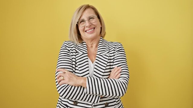 Happy midlife blonde woman confidently posing in striking striped jacket, arms crossed, beaming smile towards camera. a picture of life-affirming positivity against a cool, isolated yellow backdrop