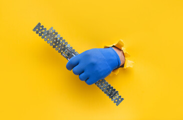 A left man's hand in a blue knitted glove holds a U-shaped fastening profile for drywall. Torn hole...