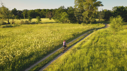 Traveler girl ride on bicycle at green grass field aerial. Summer nature landscape. Leafy forest at...