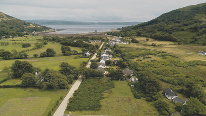 Slow motion of Scottish whisky distillery at village aerial. Road, cottages, houses at green mountain valley. Summer cloudy day. Nobody nature landscape of Arran Island, Scotland. Cinematic drone shot
