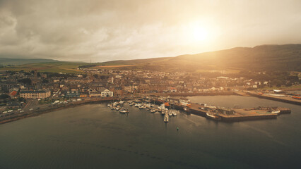 Pier town citycsape with historic buildings at ocean bay aerial. Sun shine over ships, yachts at marina wharf. Streets, road with cars, trucks. Green hill, valley at Campbeltown city, Scotland, Europe